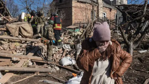 A woman surrounded by rubble covers her mouth as she cries in front of a damaged house