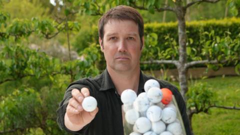 A head and shoulders image of a man wearing a dark coloured shirt while sitting in a garden with trees behind him. He is holding a white golf ball to the camera while looking into the camera. In front of him is a clear receptacle filled with golf balls - all white ones, except for one orange one.