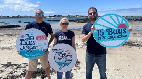 Two men and a woman in sunglasses and t-shirts stand on a white sandy beach with the sea behind them, each holding a sign promoting the 30 Bays in 30 Days events