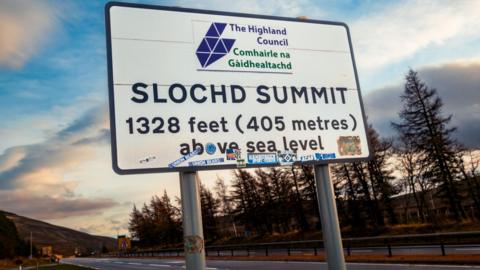 A large road sign on the A9. It has Highland Council's name in English and Gaelic and the words "Slochd Summit" and its height above sea level - 1,328ft (405m).