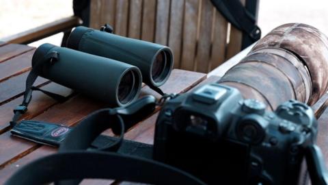 A pair of binoculars and a camera with a long lens are on a table. A chair is next to the table. 