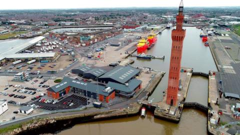 Aerial view of Grimsby Dock Tower. It is surrounded by an expanse of water, with  industrial buildings and boats in view.