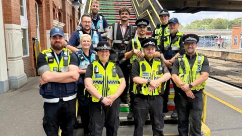 A large group of police officers stand, smiling at the camera. They all have their hands clasped in front of them and are standing on a train platform on a set of stairs. The front row of officers are wearing black uniform with yellow high vis vests and black hats. Other officers in the background are wearing navy blue uniform with blue vests, signifiying they are transport police.