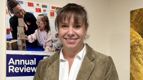 A woman wearing a white shirt under an olive blazer with a poster behind her with a child and carer playing with building blocks.