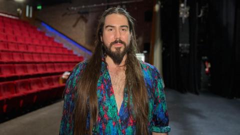 Adam looks at the camera as he wears a bright shirt which is unbuttoned down his chest. Adam has long dark brown hair which comes past his chest and a beard. Behind him is rows of theatre seats in the Arts Centre