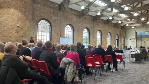 Rows of people seated on red chairs in a large room. They are facing committee members, who are seated in an oval-shaped arrangement.  