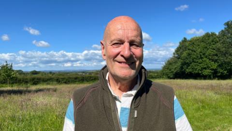 Photo of David looking into the camera and smiling. He is wearing a blue and white striped rugby shirt with a dark khaki gilet over the top. He is stood on the edge of a meadow with wild grasses - the sky is bright blue and there is plenty of sunshine.