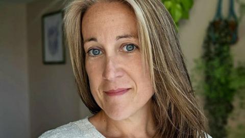 Poppy Whiteside smiling slightly at the camera with her home, with a framed piece of art and plants on the wall, in soft focus in the background