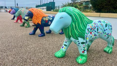 Five colourful lion sculptures lined up on a forecourt outside Cheltenham Racecourse. The nearest lion has green main and a light blue floral design on the body. The next nearest lion has an orange main and dark blue body and the others are green, purple and orange.