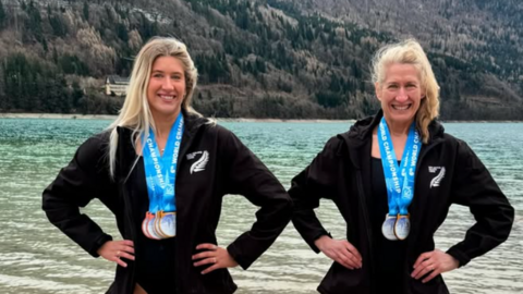 Two blonde women in black wet suits with medals around the necks stand with their hands on their hips in front of a lake smiling at the camera.