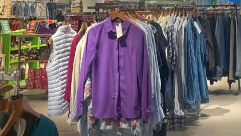 Second hand clothes including various items on rails in a shop. A purple shirt is at the front of the rack and is centre of the photo. 