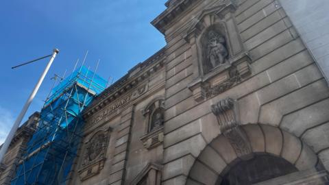 An upward view of a historic library façade featuring a stone statue in a niche, with scaffolding and blue netting to one side.
Decorative stonework and a carved figure on a civic building are seen against a bright blue sky, with refurbishment scaffolding alongside.
The upper section of a classical library building shows ornate detailing and a statue, partially framed by construction scaffolding.
A heritage building façade with carved stone features and a figure labelled Dryden, with blue netting covering nearby scaffolding.