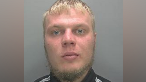 A police custody photo of Eimantas Zemeliauskas. He has short blonde hair and a wispy beard. He is wearing a black top with a white stripe over each shoulder