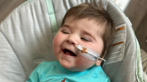 Jack has short brown hair and is wearing a turquoise top and sitting in a baby seat. He has a tube up his nose which is taped to his face with a plaster with a teddy bear on it. He is laughing.