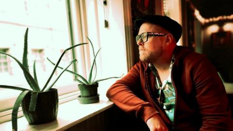 Phil Jackson sits next to a window and looks outside. He has a black cap on, glasses, a dark orange jacket and top underneath. There are two plant pots sitting on the windowsill next to him.