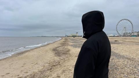 A picture of Leon in a black hoodie, with the hood covering his face to protect his identity. He is pictured on Great Yarmouth's central beach, with the Wellington Pier and big wheel visible in the distance.