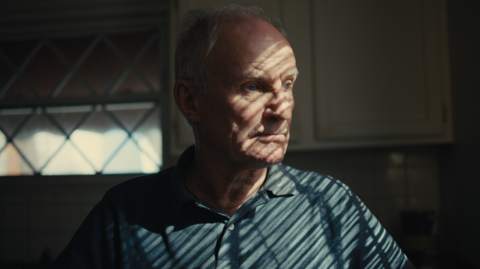 A man is looking through a window, with a long distant gaze.
