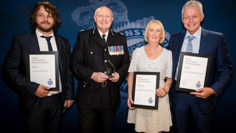 John Harrop wears a dark-coloured suit and blue tie, standing next to Chief Constable Mark Roberts who holds a glass award, with Geraldine Baddley, wearing a cream dress with a dotted design, and Phillip Baddley, wearing a blue suit and tie, stand to the right of the photo. All of the award recipients hold a framed certificate. 