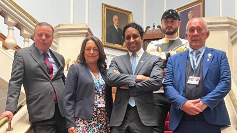 Five members of Reform, four men wearing suits and a woman in a floral dress and grey jacket, are posing on a staircase