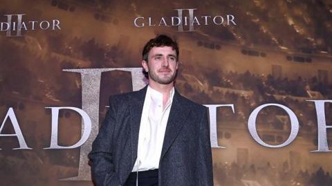 Paul Mescal who has a brown short/curly mullet standing in front of "Gladiator II" premier at Light House Cinema in Dublin.  He is wearing a white shirt and a grey jacket