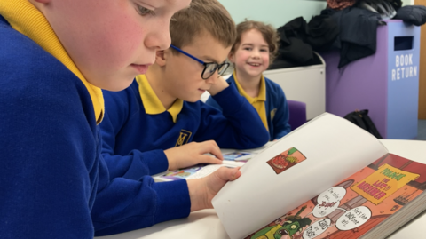three children aged 7 or 8 reading books, there are two boys and one girl and they are in a blue uniform with yellow tops