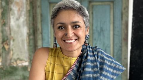 A woman smiling and looking at the camera. She is wearing large silver earrings and has a silver nose piercing. She is wearing a yellow top with a blue and white shawl draped over it. In the background, which is out of focus, there is a blue door.