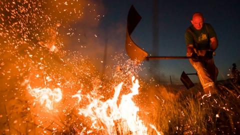 Firefighters tackle a wild fire on Winter Hill near Bolton in 2018. It is dark and the fire is a white hot blaze being batted by a fireman sending sparks into the sky