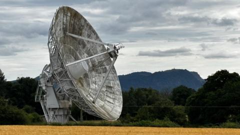 There is a big radio telescope dish which is worn. It has hints of white but is largely dirty and has darkened shades of black. It is on top of a field of yellow and has trees in the background.