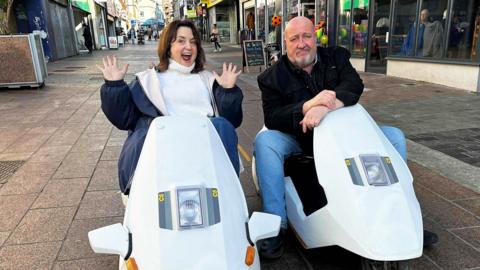 Actors Ruth Jones and Steve Speirs sat in retro Sinclair C5s in a street filled with shops