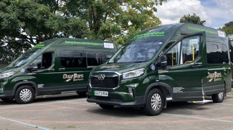 Two green minibuses with OurBus Bartons written on them. They are parked on grey tarmac.