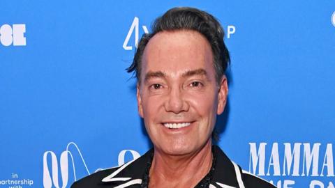 Craig Revel Horwood standing in front of a blue banner smiling at the camera. He has black hair swept to one side and is wearing a black top with a white trim on the collar.