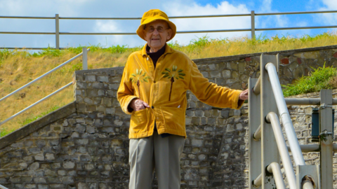 Maurice is posing for his photograph outdoors and wears a bright yellow jacket with two flowers on and a matching yellow bucket hat.