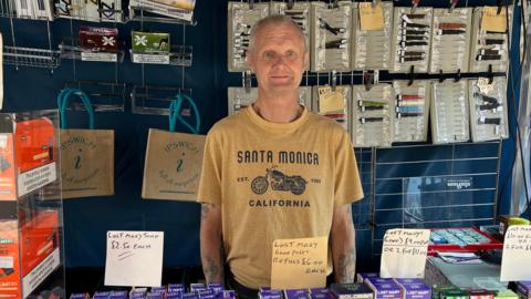 A man with grey hair wearing a yellow T-shirt stands inside a stall. There are vape products hanging on a blue wall behind him as well as under items lying on a table in front of him.