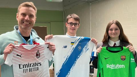 Vice Principal at the academy, Michael Cox, with pupils Jack and Amelia. They are stood side by side and each holding a football shirt up in front of them. There is an AC Milan, FC Dynamo Kyiv and FC Barcelona shirt. 