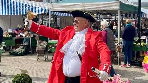A man wearing a black tricorn hat and red jacket over a white shirt with lace ruff, is ringing a bell in a town centre. It appears to be during a market day.