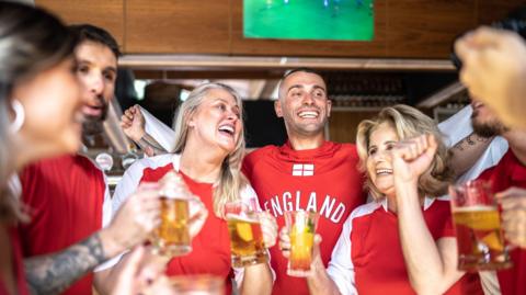 A stock image showing a group of six people wearing red England shirts, drinking pints of beer in a pub, while a TV screen plays the football match in the background.