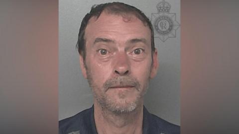 A police head and shoulders photo of a man with short brown hair and grey stubble looking directly into the camera. He is wearing a black top and the background is grey.