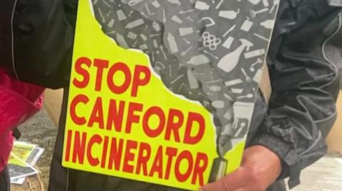 A person holding a placard with the words 'Stop Canford Incinerator' in red text