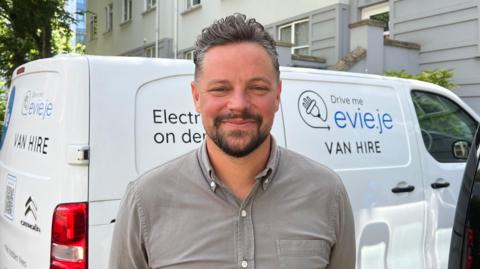 Nick Perchard-Rees facing the camera infront of an EVie electric van. He has short greying hair, and dark beard and is wearing a grey button-up shirt.