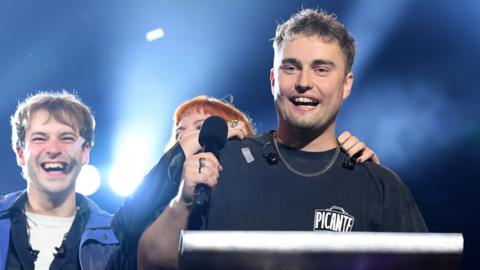 Sam Fender holds a microphone onstage while smiling alongside two of his band members who are also looking joyful, backlit by the stage lighting, at the Mercury prize ceremony in Newcastle on Thursday evening.