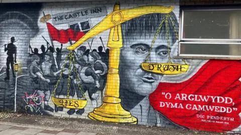 A mural painted onto an outdoor brick wall in Port Talbot, the artwork is a memorial to Dic Penderyn (Richard Lewis), A large, yellow scale of justice is unbalanced, labeled "LIES" weighing heavily downward on the left, while the pan labeled "TRUTH" elevated on the right. With a painting of Richard Lewis' face behind the scales.