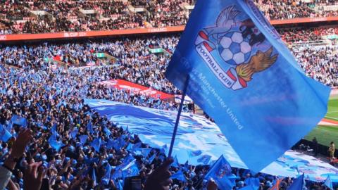 Sky Blue Flag with the crest of Coventry City is waved in the foreground with a background showing the stands at Wembley stadium filled with thousands of Sky Blue shirts.