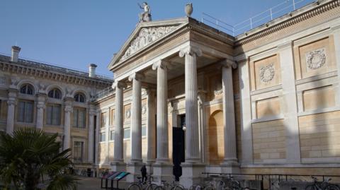 Outside the Ashmolean Museum. There are several bikes at its bike racks. A person is seen approaching the entrance. It is sunny.