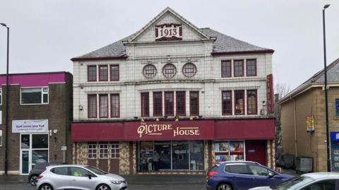 The ornate cream and maroon exterior of a 1913 cinema with cars driving past its front entrance and gold lettering spelling out The Picture House