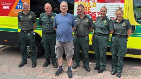 Jason Pizzey stood in front of the ambulance staff that saved his life and an amblance is parked behind them