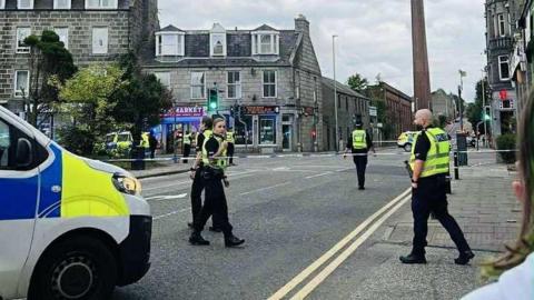 A group of police officers, all wearing black uniforms and hi-vis vests, walking behind a police cordon on a street with shops.