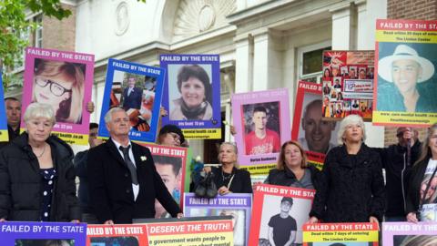 The families of those who have died while in the care of mental health services in Essex stand outside the inquiry protesting. There are around 20 people in the image which shows them standing with placards held against them and large posters of their loved ones. They show a range of faces, both men and women and young and old. One reads "failed by the state".