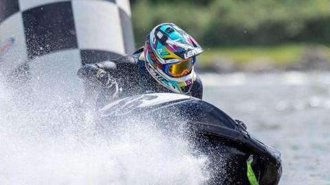 A person rides on a jet ski with a colourful helmet with water spraying towards the camera