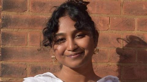 A portrait of a woman standing outdoors against a red brick wall. She is wearing a loose, white button-up shirt and small hoop earrings. Her hair is dark and styled up, with loose strands framing the face. 