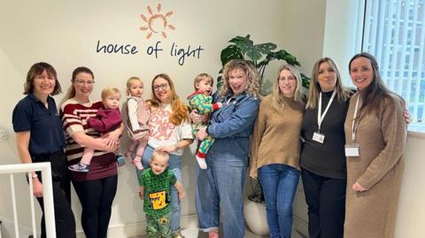 A group of women - mums with babies and toddlers and employees of the charity - all stand together a smile for a group photo.
